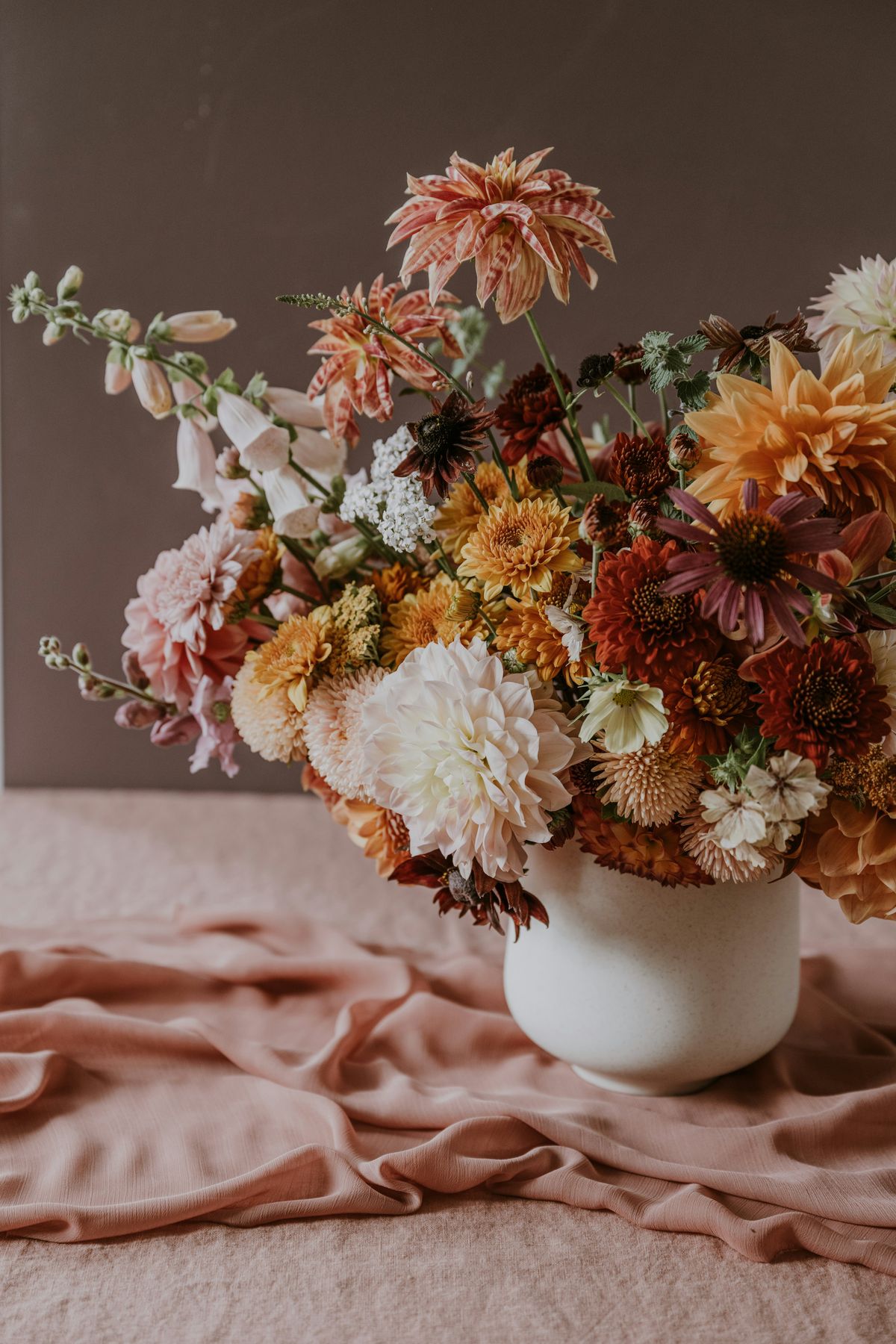 A loose arrangement of dahlias in peach, rust and cream in a white vase on blush linen